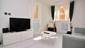 Ett modernt vardagsrum med en platt-TV placerad på ett vitt skåp, ett runt soffbord, en grå soffa och en flätad fåtölj med en kudde.