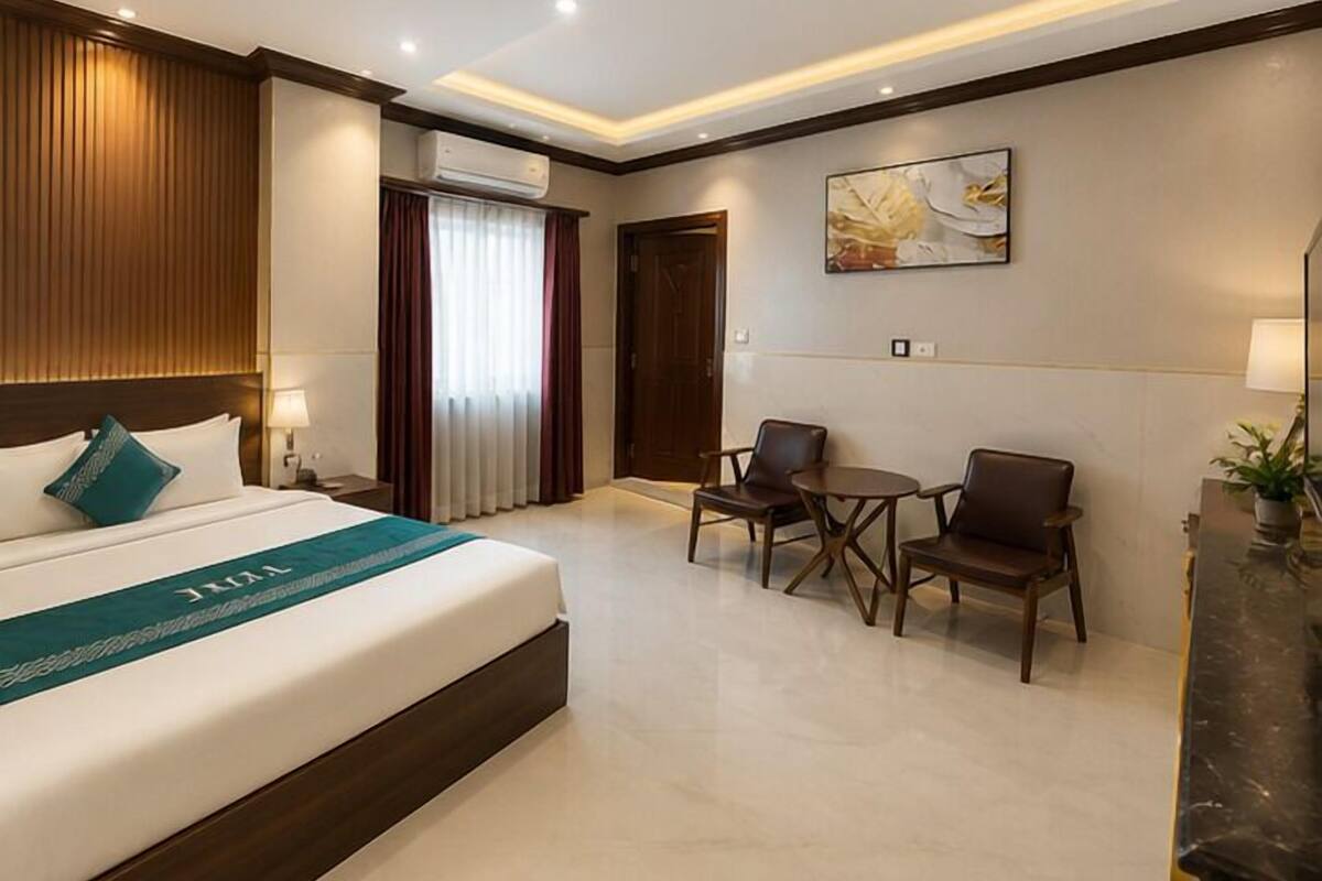 A hotel room with a bed, a desk, two chairs, a window with curtains, an air conditioning unit, and a painting on the wall.