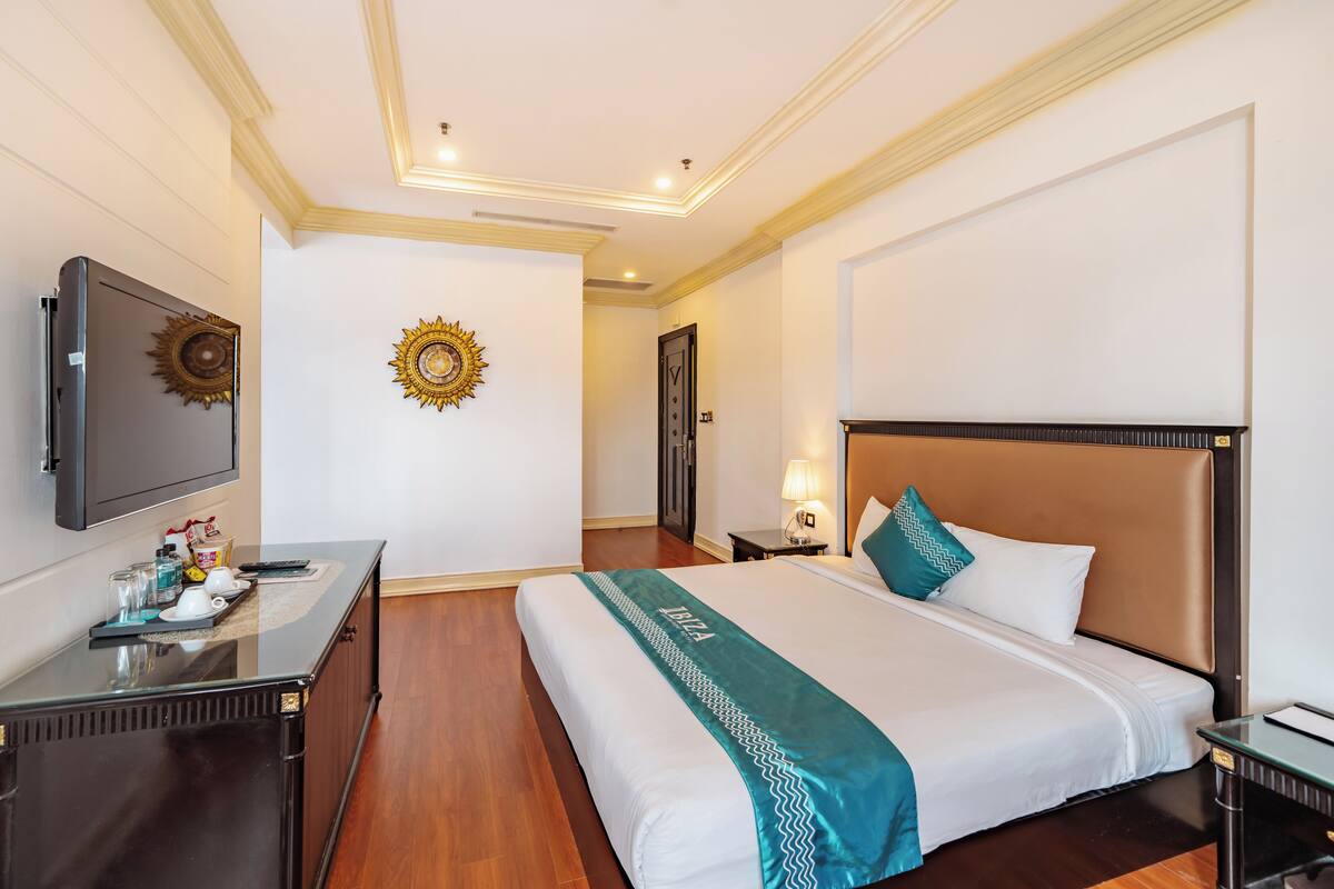 A hotel room with a large bed, a desk with a flat-screen TV, a nightstand, and a wall-mounted sunburst decoration.
