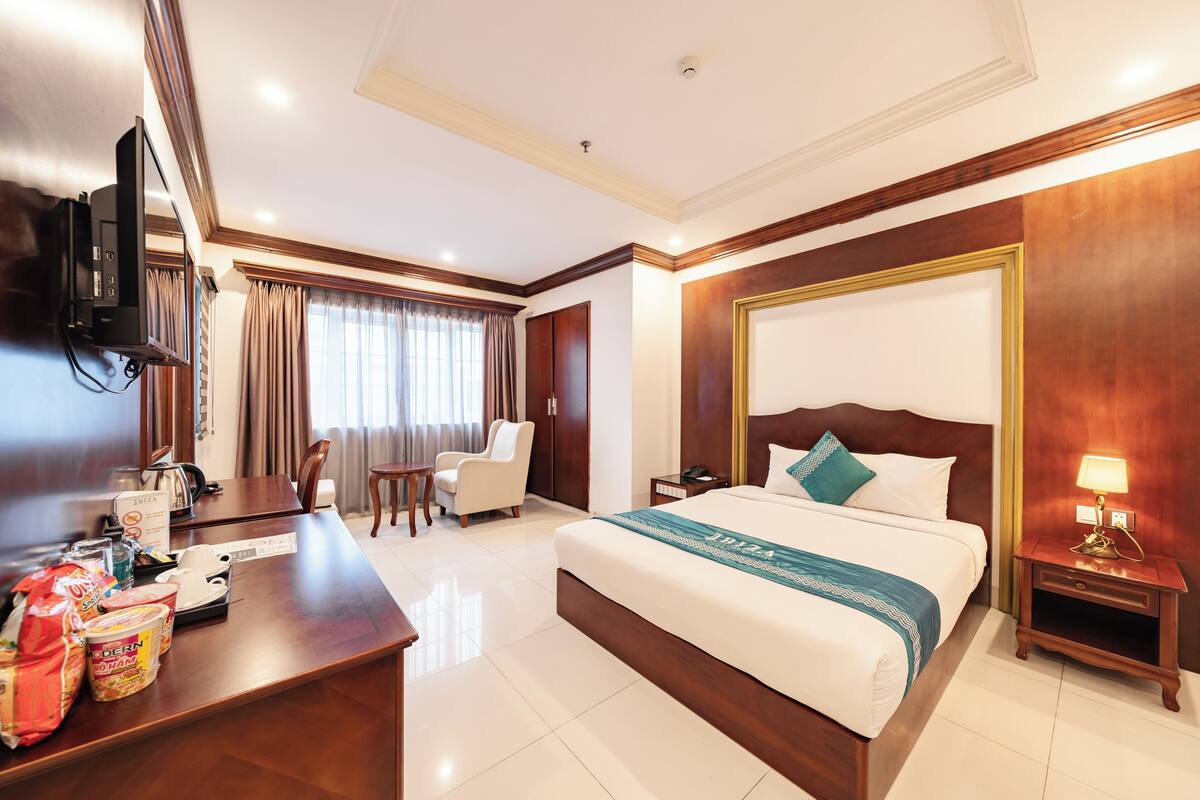 A hotel room with a bed, a desk, a chair, a TV, and a nightstand with a lamp.