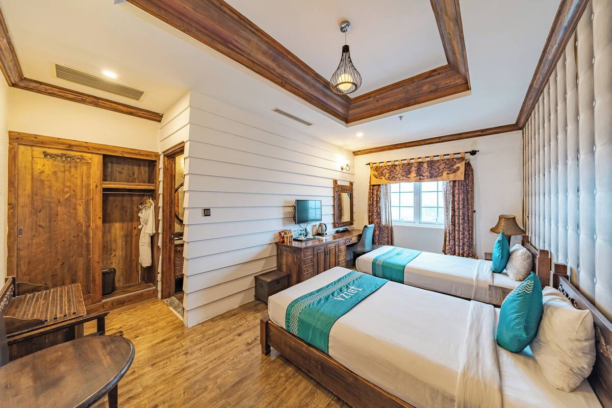 A hotel room with two beds, wooden furniture, a desk, a mirror, and a window with curtains.