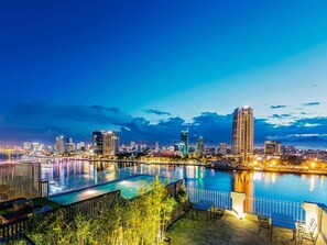 A nighttime cityscape with a large, illuminated swimming pool and a view of the city skyline. - IBIZA Riverfront Da Nang Hotel (Da Nang)