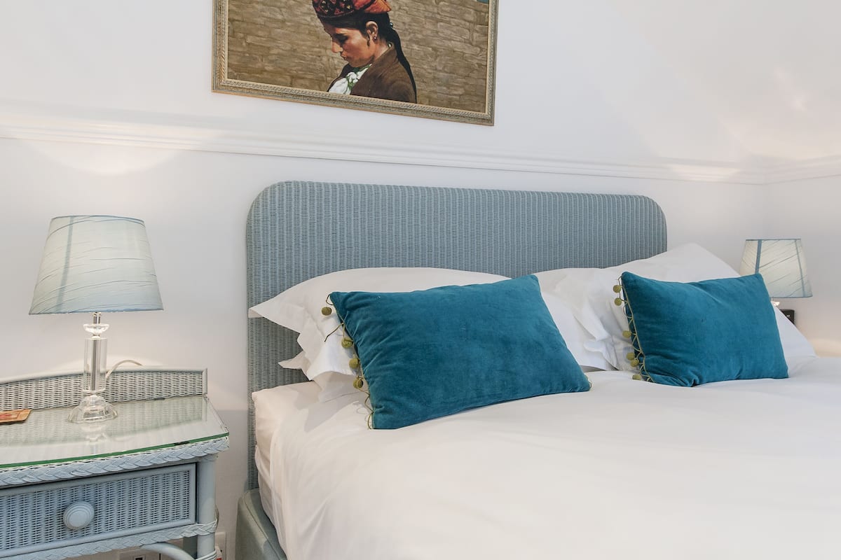 A neatly made bed with white and teal pillows, a glass side table with a lamp, and a framed picture on the wall.