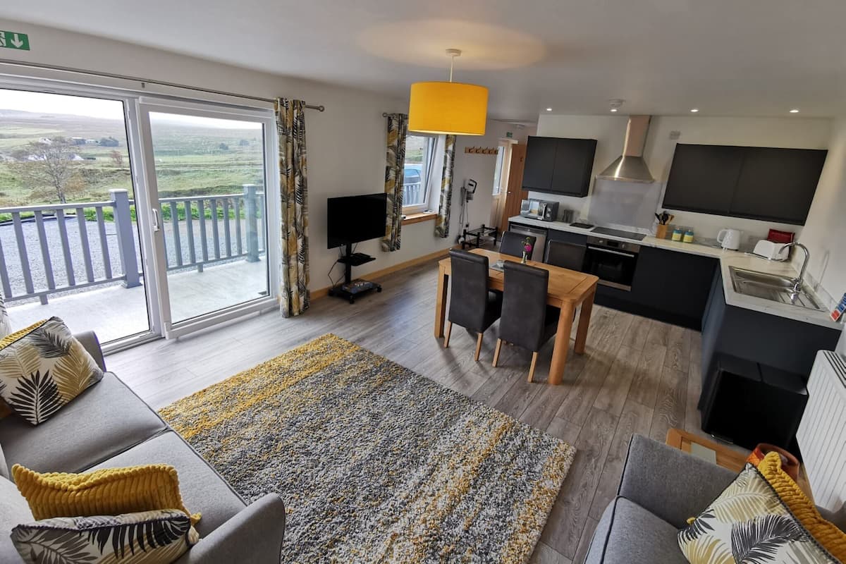 A modern living room with a sofa, a dining table, and a kitchen area with a TV and a yellow pendant light.