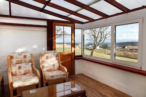 Una habitación con dos sillas de mimbre, una mesa de madera y un amplio ventanal con vistas a un paisaje cubierto de hierba y colinas al lejano.