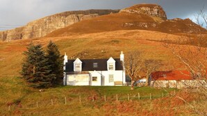 Une maison blanche avec un toit noir, entourée d’un champ clôturé, et une grande colline en arrière-plan.