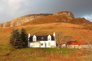 A white house with a black roof, surrounded by a fenced field, and a large hill in the background.