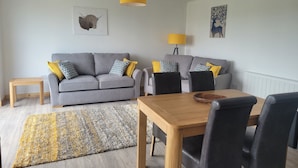 Un salon avec un canapé gris, une table à manger en bois et des chaises noires. On y trouve deux lampes jaunes, un tapis à motifs et des œuvres d’art accrochées aux murs.
