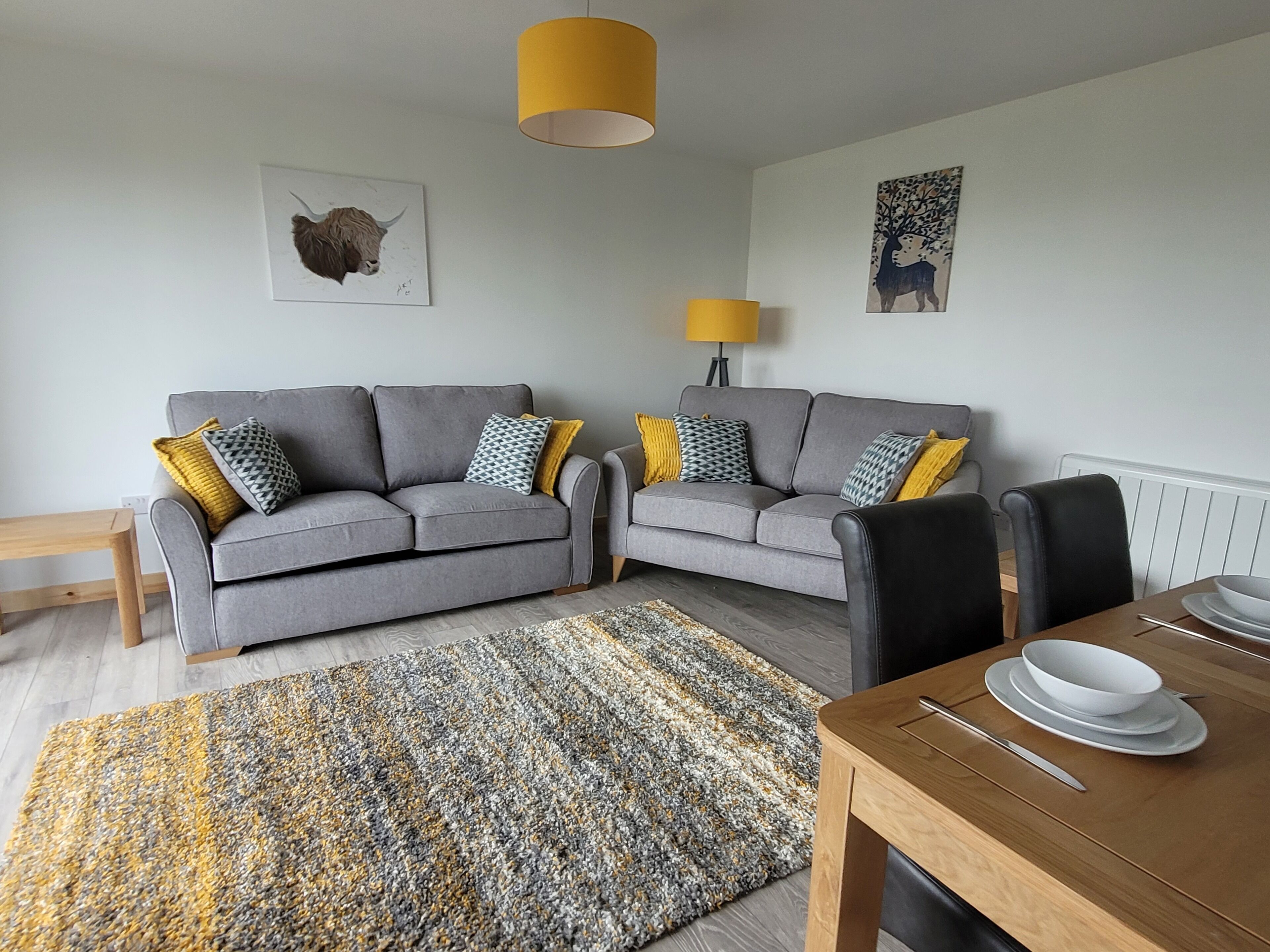 A modern living room with two grey sofas, a wooden dining table with white plates, a patterned rug, and two yellow lamps.