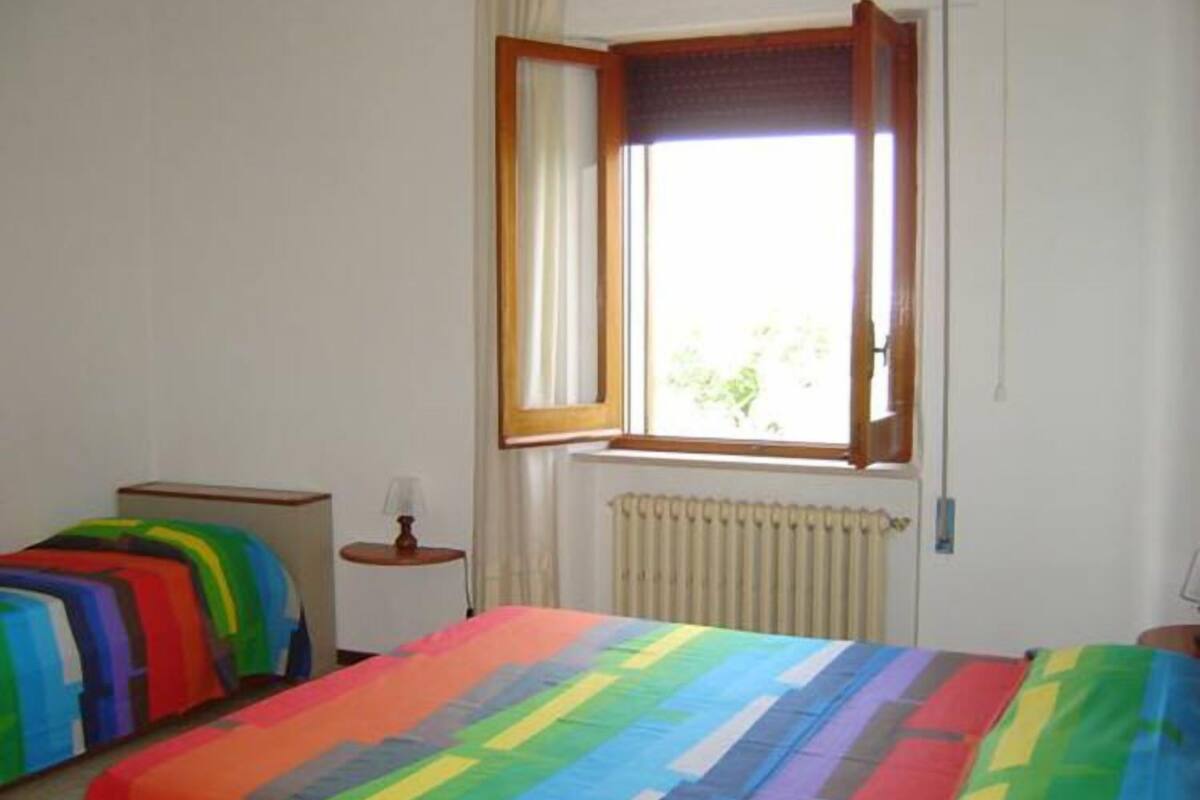 A room with two beds, a window with curtains, a radiator, and a small table with a lamp.