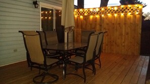 Relax on the outdoor patio with a dining table and four chairs, surrounded by a wooden fence and string lights.