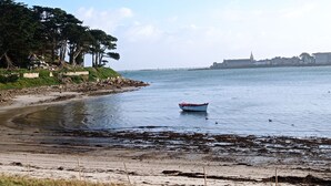 Eine Küstenansicht mit einem kleinen Boot auf ruhigem Wasser, einem sandigen Strand mit Seetang und einer in der Ferne liegenden Stadtlandschaft mit einem markanten Gebäude.