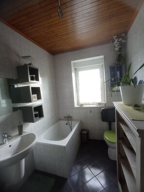 Bagno con vasca, water, lavabo, soffitto in legno e una finestra con una pianta.