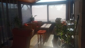 Ein sonnendurchflutetes Zimmer mit Korbmöbeln, Holzbalkendecke und großen Fenstern.