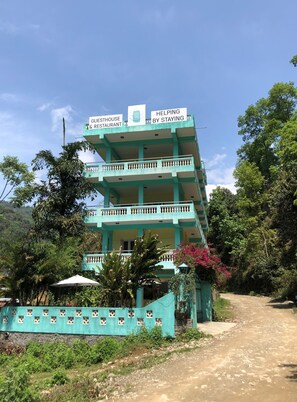 Un edificio de varios pisos con balcones, identificado como casa de huéspedes y restaurante, rodeado de vegetación.