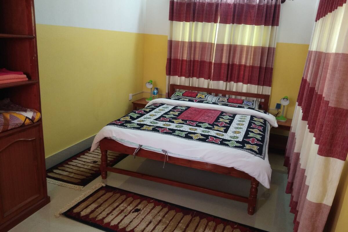 A bedroom with a wooden bed, a bedside table with a lamp, a shelf with folded clothes, and a window with curtains.