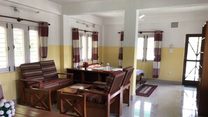 A room with a sofa, chairs, a table, and a bed. There are windows with curtains, a ceiling fan, and a door. - Happy Sedi Lodge (Pokhara)