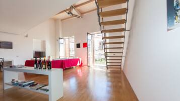 A spacious room with a wooden floor, a white desk with books and wine bottles, a red sofa, and a staircase.