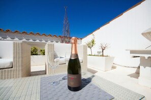 A bottle of wine on a table in an outdoor seating area with white wicker furniture and a clear blue sky in the background.
