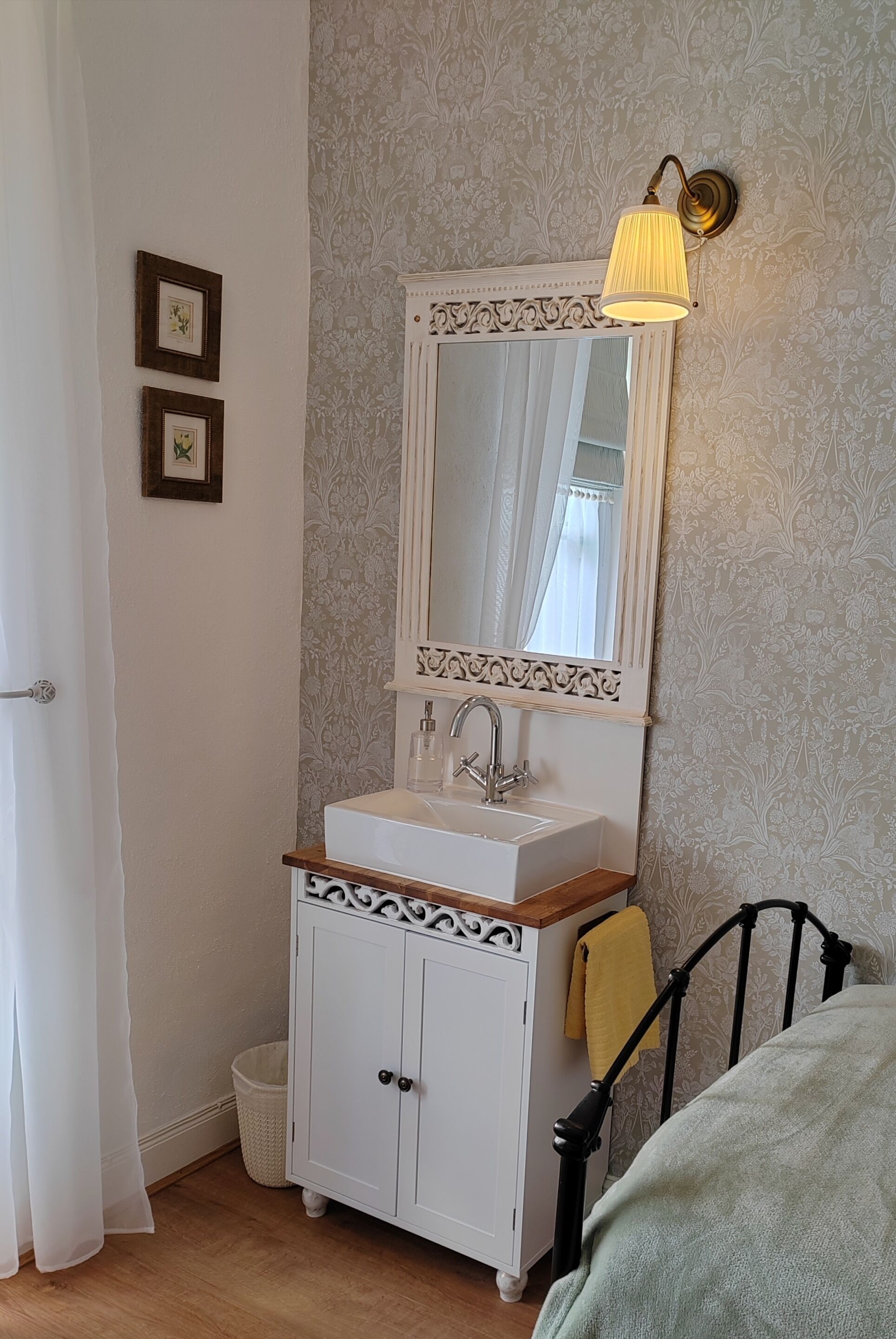 A bathroom with a white vanity, a mirror with a decorative frame, a wall-mounted light fixture, and a bed with a yellow towel.