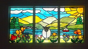 A stained glass window with a pastoral scene, including a sheep, flowers, and a sailboat. - Puddle Duck Lodge (Windermere)