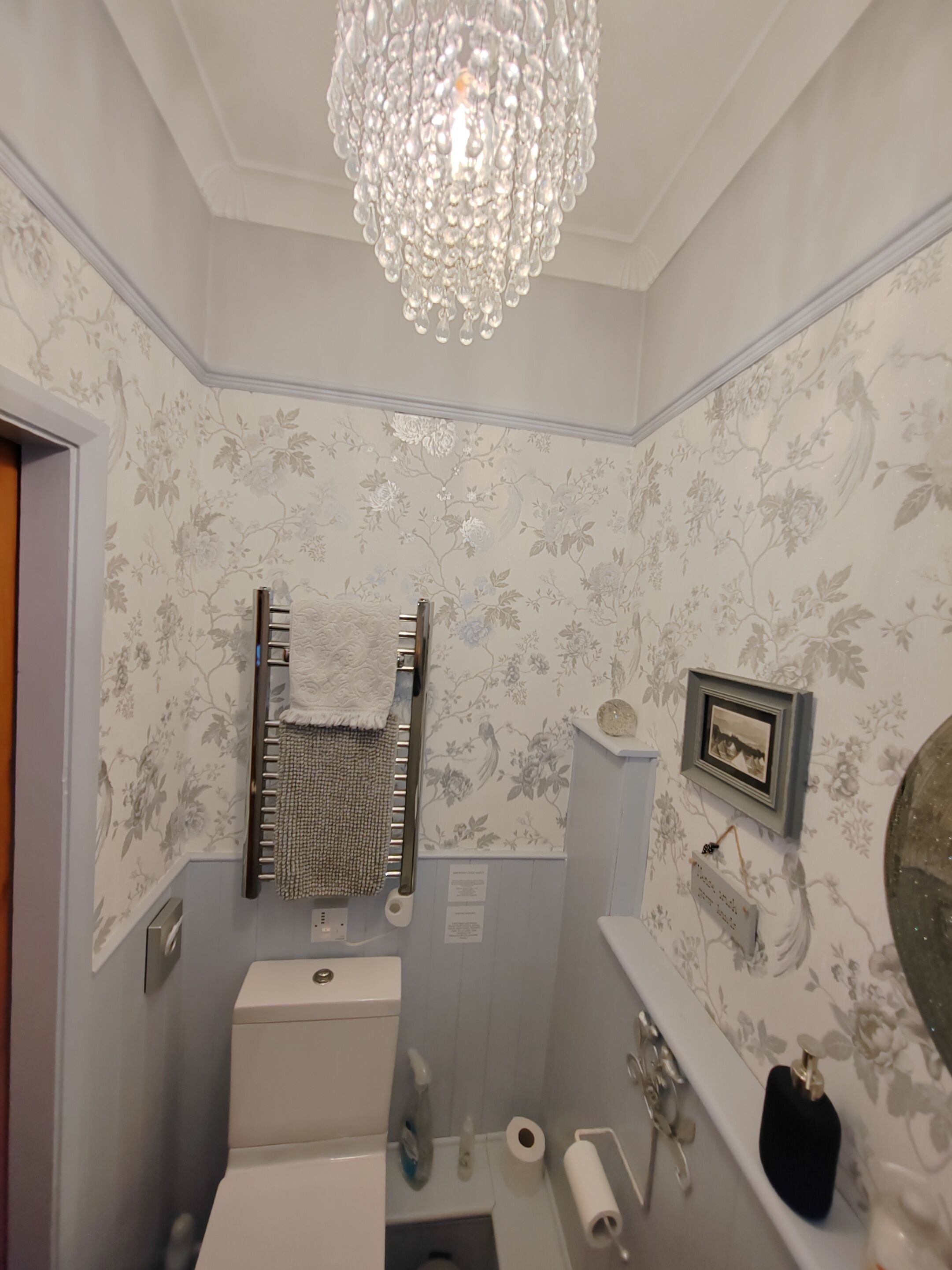 A bathroom with a toilet, sink, and a shower. There is a chandelier above the sink and a framed picture on the wall.