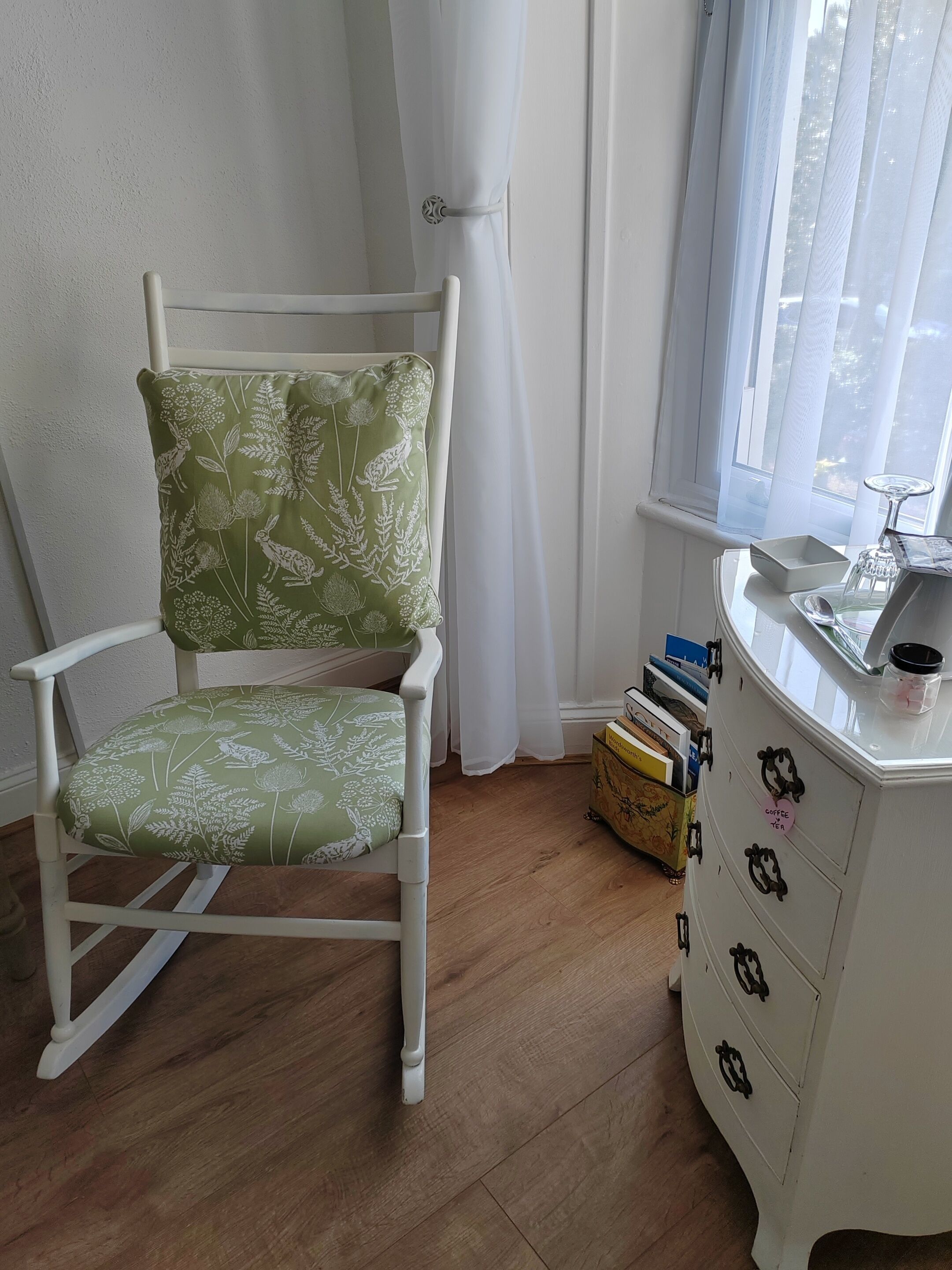 Une pièce comprenant un fauteuil à bascule blanc avec un revêtement floral vert, une commode blanche avec des poignées noires et une fenêtre ornée de rideaux blancs et légers.