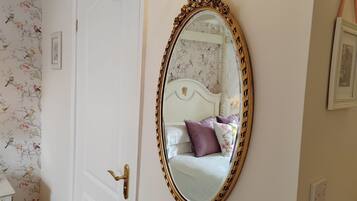 Ein Schlafzimmer mit weißer Tür, einem großen, verzierten Spiegel, blumigem Tapetenmuster und einem Bett mit violetten Kissen.
