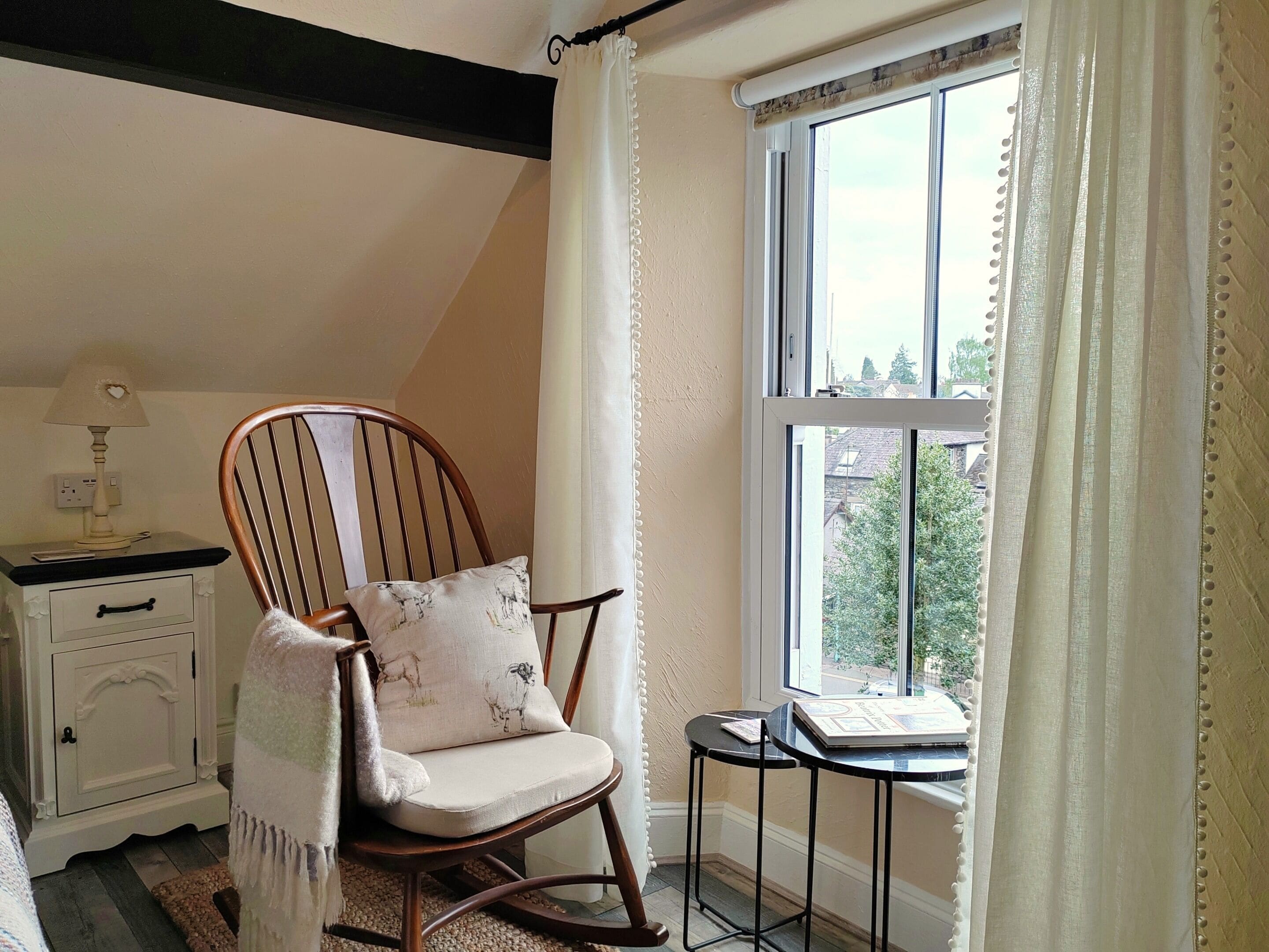 Ein gemütliches Zimmer mit einem Holzschaukelstuhl, einem kleinen Tisch und einem fenster mit Vorhängen.