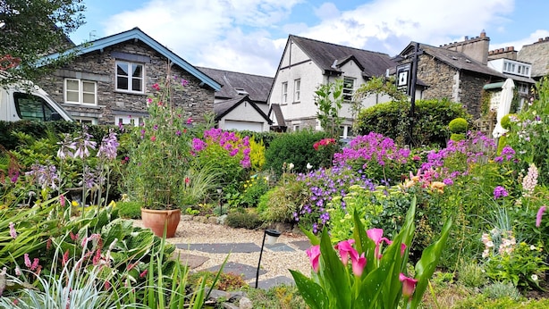 A garden with various plants and flowers, a stone house with a blue roof, and a pond. - Puddle Duck Lodge (Windermere)