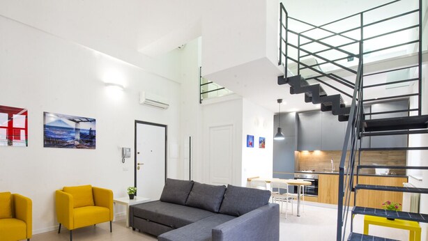 Una sala de estar moderna con un sofá gris, dos sillones amarillos y una escalera que lleva a una zona de cocina.