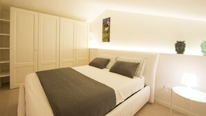 Un dormitorio con cama, mesita de noche, lámpara, estante en la pared con adornos y un armario grande.