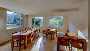 A spacious dining area with wooden tables and chairs, large windows offering views of greenery and mountains. - Blossom Bliss Resort (Dhari)