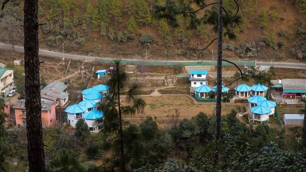 An aerial view of a rural area with several buildings, including a house with a blue roof and a white building with a blue roof, surrounded by trees and a road. - Blossom Bliss Resort (Dhari)
