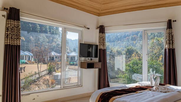 A bedroom with a bed, a TV, and large windows with curtains. - Blossom Bliss Resort (Dhari)