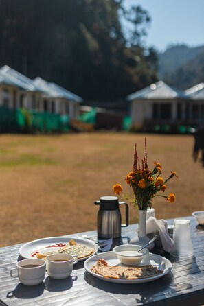 Una mesa al aire libre con platos de comida, vasos, un florero con flores y una tetera.