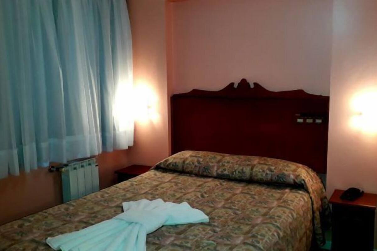 A bedroom with a bed, two bedside tables, a nightstand, a radiator, and a window with curtains.