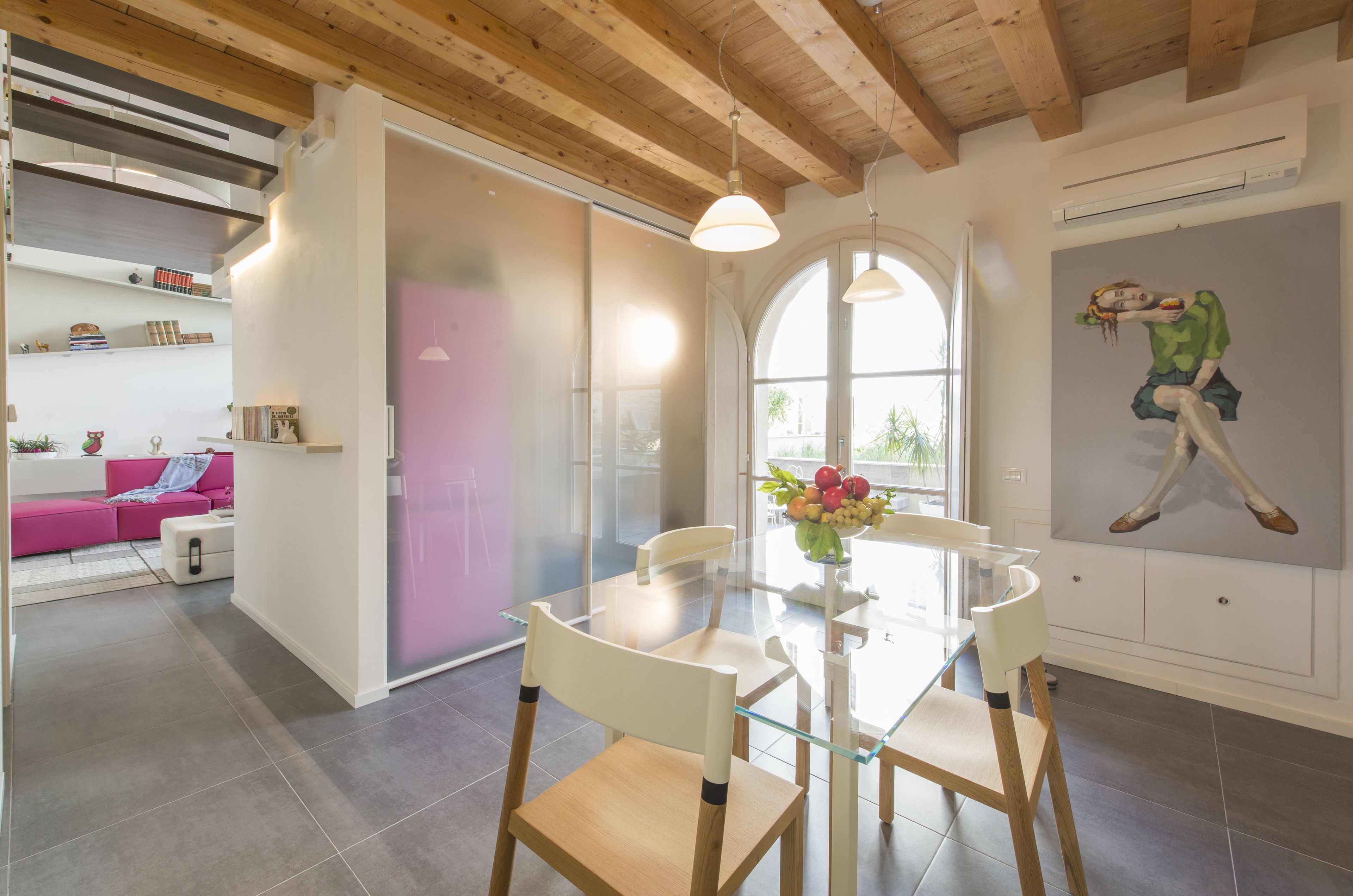 Un comedor moderno con una mesa y sillas de vidrio, un sofá rosa al fondo y un ventanal con luz natural.
