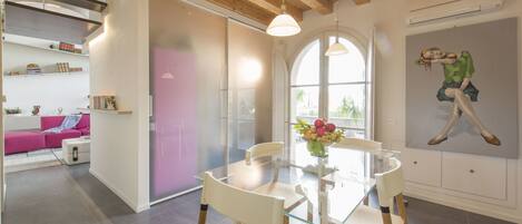Un comedor moderno con una mesa y sillas de vidrio, un sofá rosa al fondo y un ventanal con luz natural.