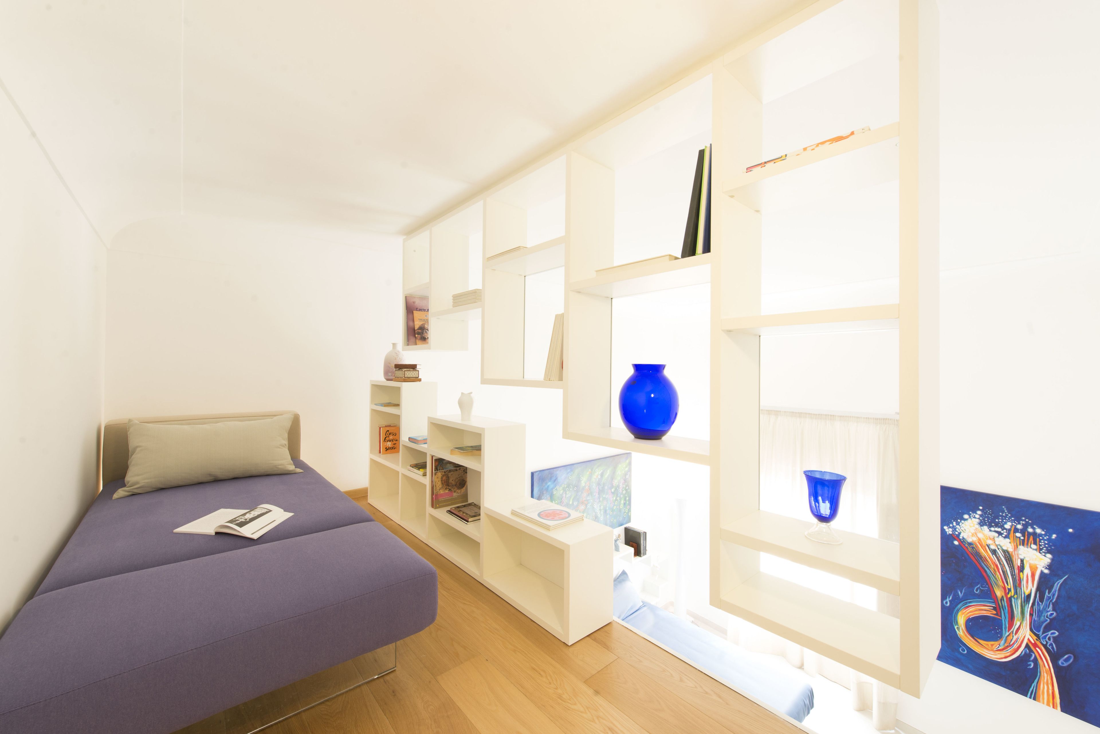 A compact room with a bed, a desk, and a shelving unit with books and decorative items.