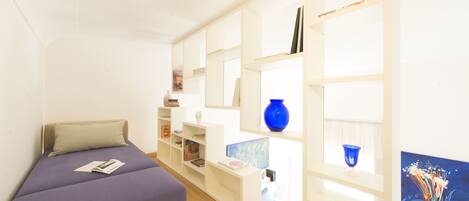 A compact room with a bed, a desk, and a shelving unit with books and decorative items.