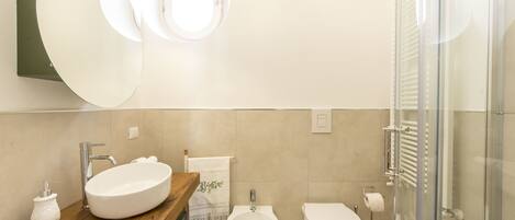 A modern bathroom with a wooden vanity, a large oval sink, a bidet, a toilet, and a glass shower enclosure.