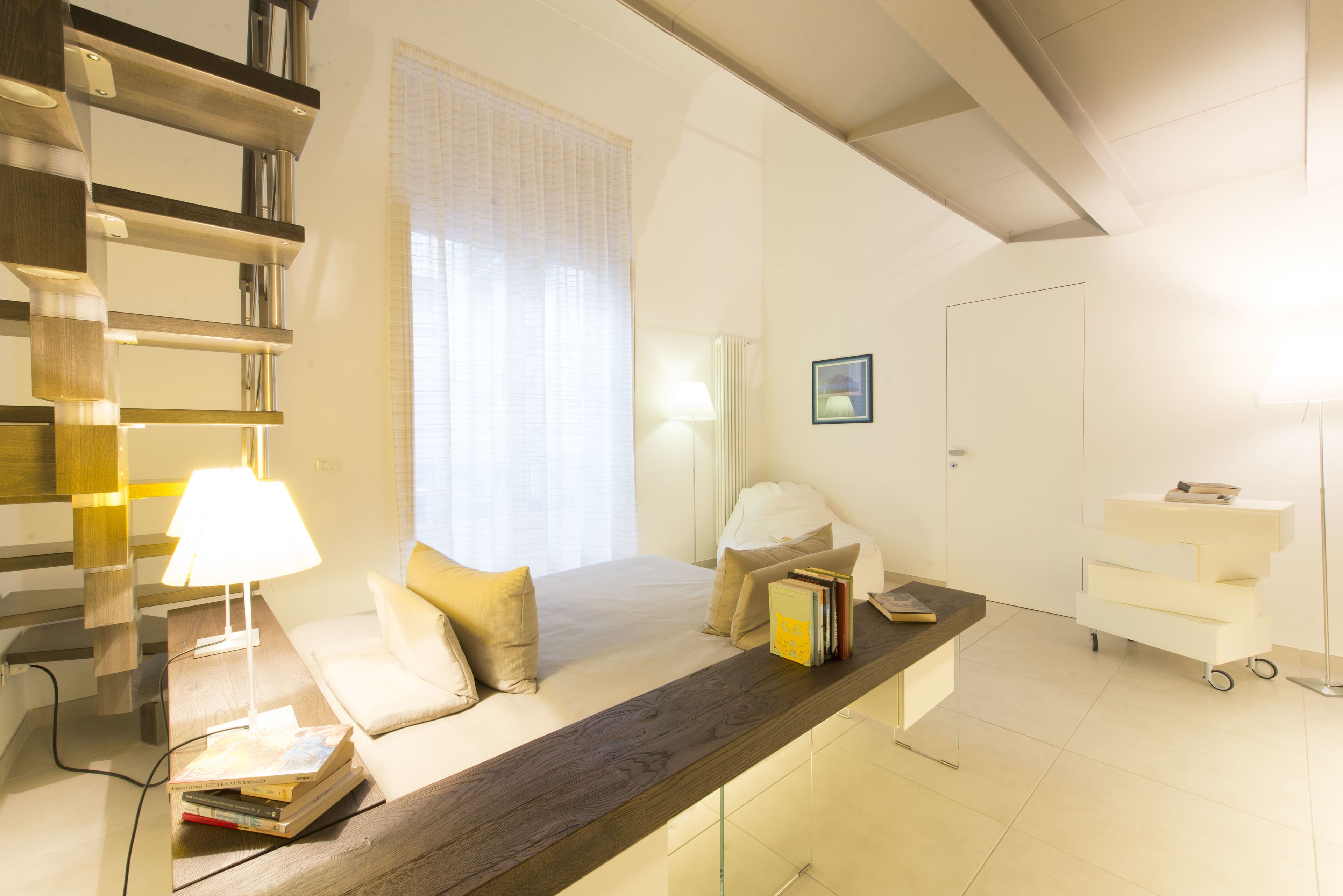 A modern hotel room with a sofa, a bedside table with books, a lamp, and a view of the outdoors through a window.