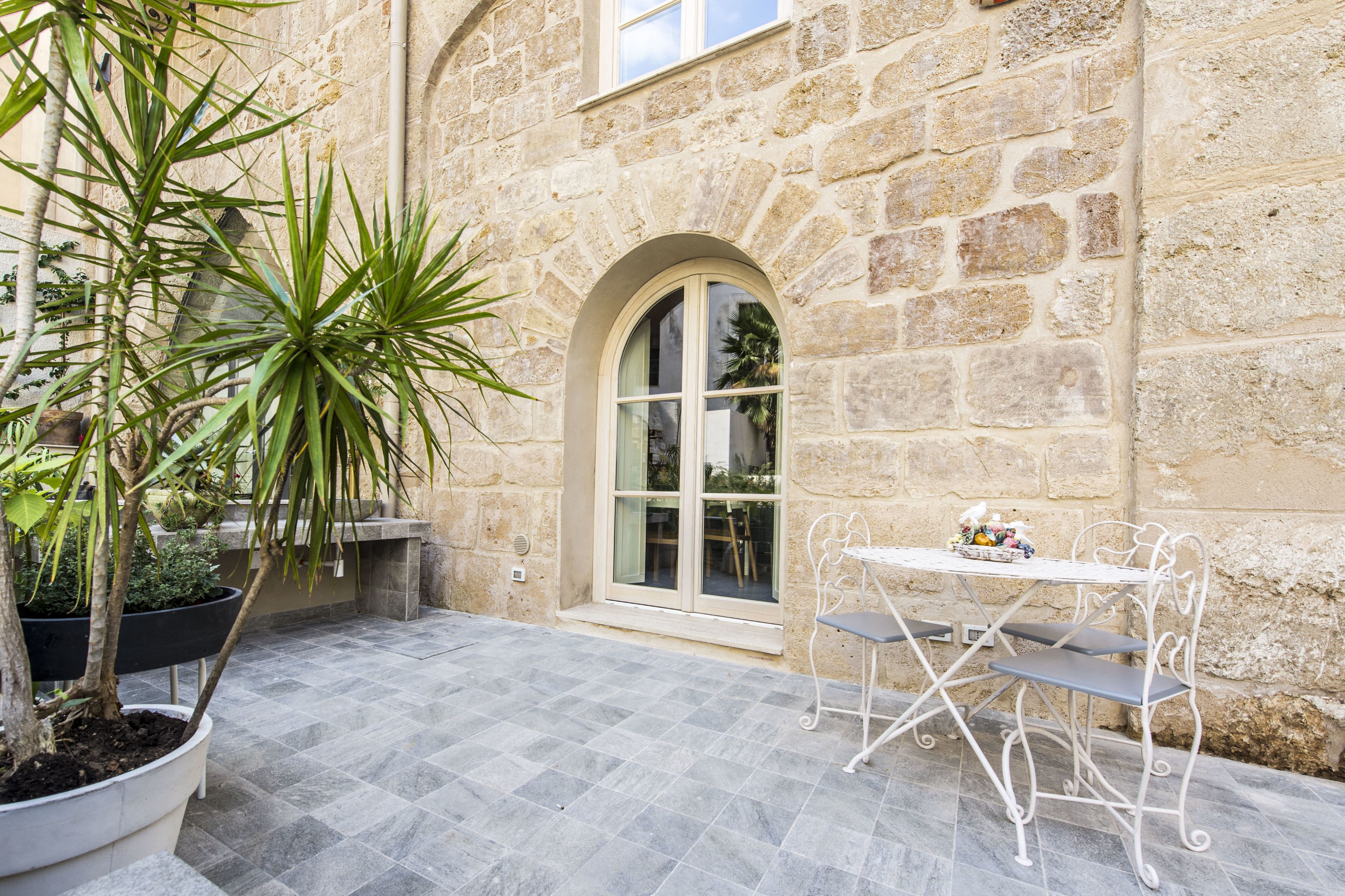 A stone building with a tiled patio, a white table with metal chairs, and potted plants.