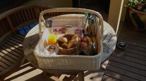 A wicker basket on a wooden deck containing breakfast items like bread, fruit, and a bottle of juice. - Schaeferwagenhotel-Campsite (Sulzfeld)