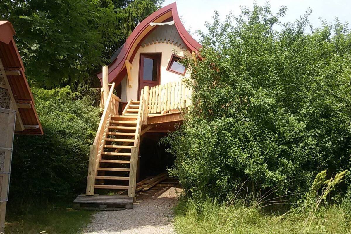 A wooden cabin with a sloped roof, a wooden staircase leading to a door, surrounded by dense greenery.