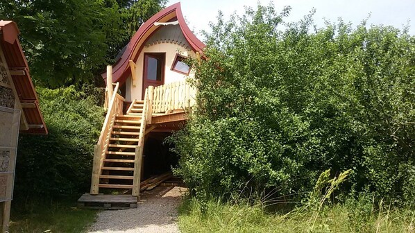 A wooden cabin with a sloped roof, a wooden staircase leading to a door, surrounded by dense greenery. - Schaeferwagenhotel-Campsite (Sulzfeld)