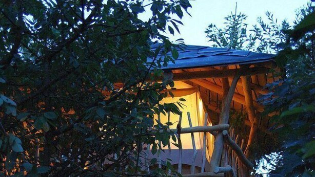 A treehouse with a wooden staircase leading up to a platform with a roof. - Schaeferwagenhotel-Campsite (Sulzfeld)