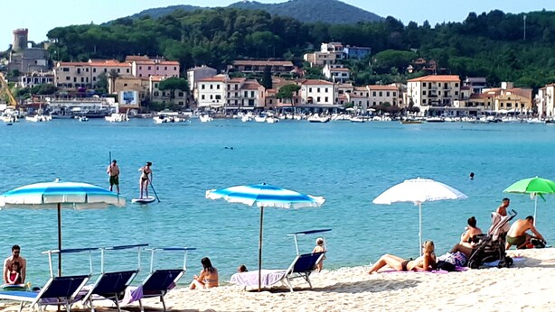 A beach with sun loungers and umbrellas, a clear blue sea, and buildings in the background.