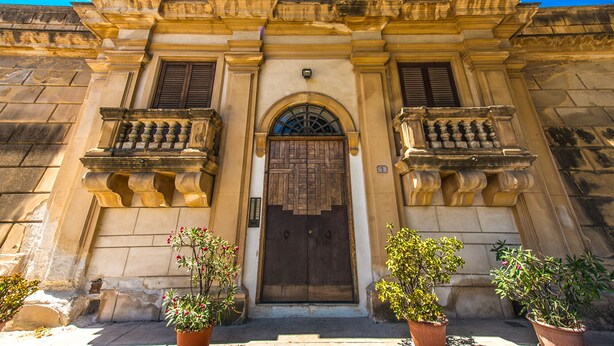 A historic building with a central arched doorway, flanked by two balconies with wrought iron railings, and potted plants at the entrance. - Amazing Sea View in Historic Building in Downtown by Wonderful Italy (Palermo)
